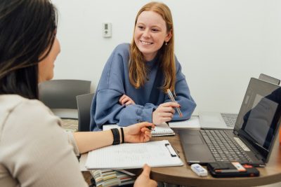 Two female college students talk to each other while sitting at a table with a laptop and notebooks.