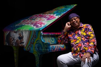 Cuban jazz pianist Chucho Valdes, an older Hispanic man, wears a purple hat and a multicolored floral button down shirt. He sits at an open grand piano covered from top to bottom in spray paint art and smiles towards the camera.