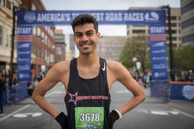 A man in running attire stands in front of a race starting line smiling.