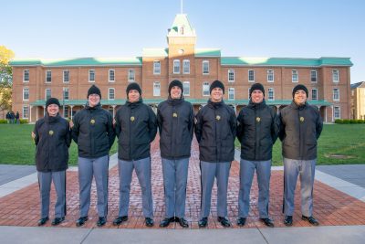 The seven cadets are all in uniform with black jackets and black hats on Upper Quad with a historic brick building in the background. 