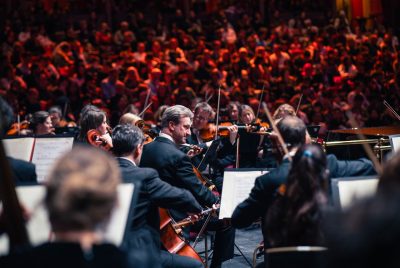 The Royal Philharmonic Orchestra performs on stage in front of a large, warmly lit audience. Musicians in formal black attire play string instruments, with sheet music visible on stands. The focus is on a cellist at the center, with other players surrounding him in close formation. The crowd in the background appears engaged, creating a vibrant, immersive atmosphere.