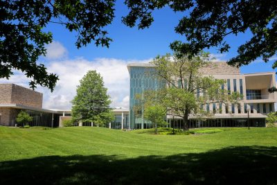 The Center for the Arts building at Virginia Tech viewed from the lawn. It's a modern building with large glass windows and a honeycomb-like facade sits on a gently sloping green lawn, surrounded by mature trees and under a bright blue sky with scattered clouds. The scene is framed by tree branches and leaves in the foreground.